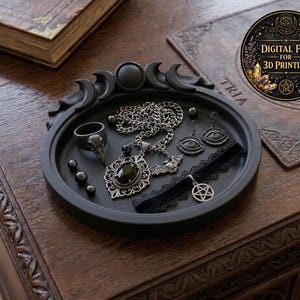 May include: A black decorative tray with a crescent moon design holds gothic jewelry. The jewelry includes a skull ring, a necklace with a large pendant, a bat pendant, eye earrings, and a pentagram choker. The tray is on a wooden surface.