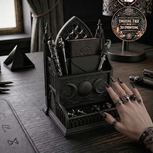 May include: A black gothic desk tidy with pen holders, a wallet, and a small drawer. The organiser features crescent moon and pentagram designs. The image also includes a hand with rings and a bracelet, and a digital file advertisement.
