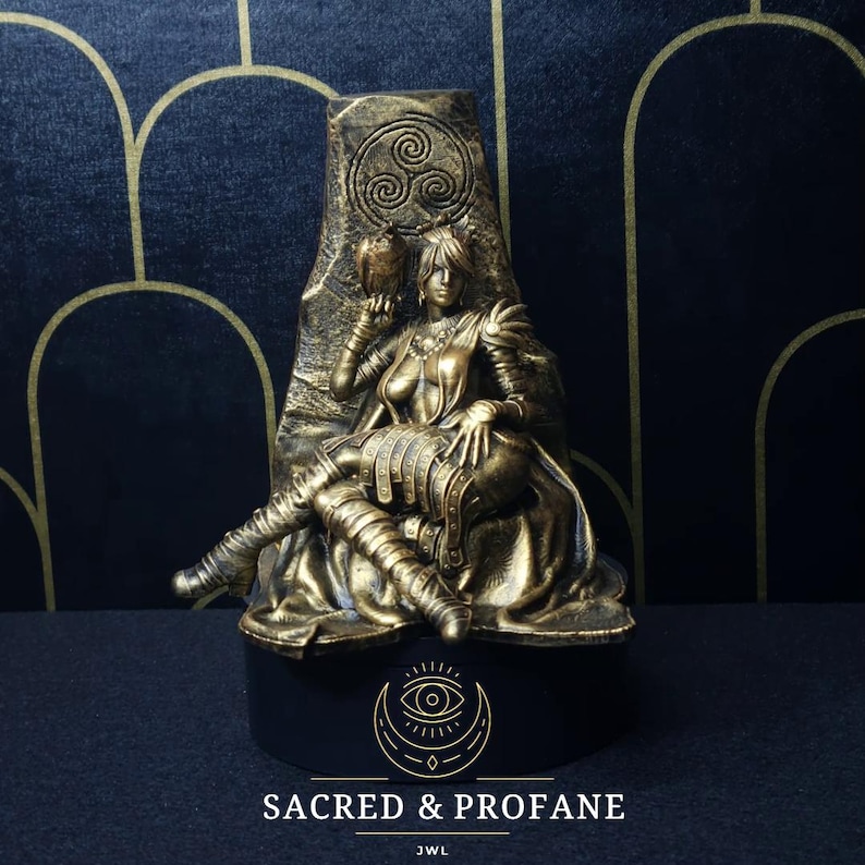 May include: A gold-toned figurine of a woman sitting on a throne. She is holding a bird in her left hand and has a crown on her head. The throne is decorated with a Celtic knot design. The base of the figurine is black and has the words "Sacred & Profane" and "JWL" written on it.
