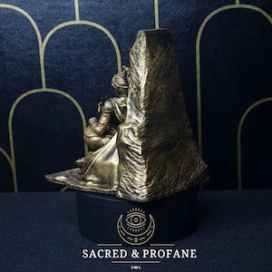 May include: A gold-painted figurine of a person sitting on a throne. The throne is made of rock and has a rough texture. The figurine is wearing a long robe and has a crown on their head. The base of the figurine is black and has a logo that says "Sacred & Profane" and "JWL".