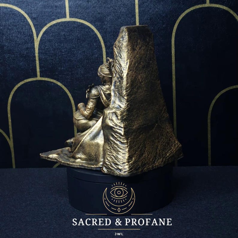 May include: A gold-painted figurine of a person sitting on a throne. The throne is made of rock and has a rough texture. The figurine is wearing a long robe and has a crown on their head. The base of the figurine is black and has a logo that says "Sacred & Profane" and "JWL".