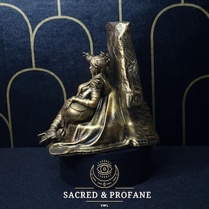 May include: A gold-colored figurine of a woman sitting on a throne. The throne is made of stone and has a carved design. The figurine is on a black base with the words "Sacred & Profane" and "JWL" written on it.