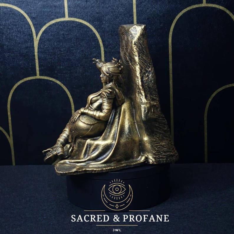 May include: A gold-colored figurine of a woman sitting on a throne. The throne is made of stone and has a carved design. The figurine is on a black base with the words "Sacred & Profane" and "JWL" written on it.