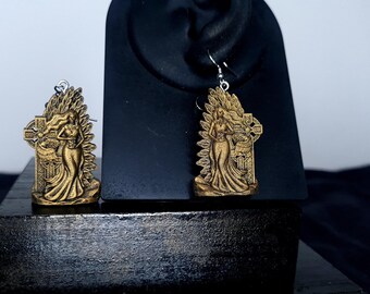May include: A pair of gold-toned earrings featuring a woman standing next to a cross. The earrings are made of metal and have a detailed design.