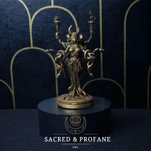 Può includere: Una statua dorata di una donna con più braccia che tengono delle torce. La donna è circondata da serpenti e si trova su una base circolare. La statua è su un piedistallo nero con il testo "SACRED & PROFANE" e "JWL" stampato su di esso.