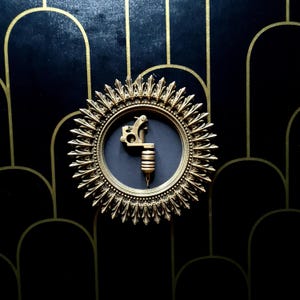 Può includere: Una cornice dorata a forma di sole con uno sfondo nero e una macchina da tatuaggio vintage all'interno. La cornice è appesa a una parete nera e dorata.