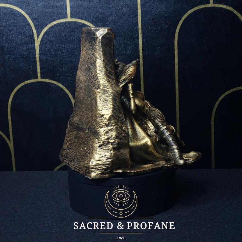 May include: A gold-colored statue of a figure sitting on a throne. The statue is on a black base with the text "SACRED & PROFANE" and "JWL" printed on it.