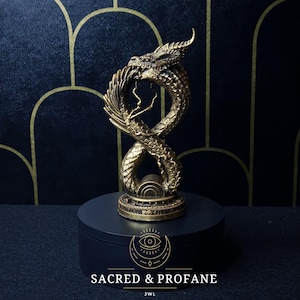 Könnte beinhalten: Goldfarbene Drachenskulptur in Form eines Unendlichkeitszeichens auf einem schwarzen runden Sockel. Der Sockel trägt den Text "SACRED & PROFANE" und "JWL".
