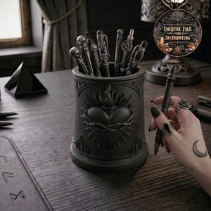 May include: A black pen holder with a Sacred Heart design, filled with various pens featuring skull and ornate designs. A hand holds a pen with a skull on top. A digital file for 3D printing is visible.
