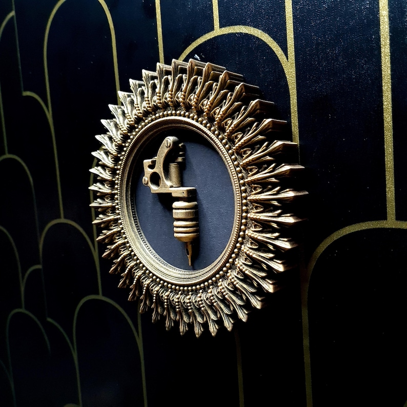 Pode incluir: Uma placa de parede decorativa dourada com uma moldura detalhada e ornamentada. A placa apresenta um fundo preto com uma m&aacute;quina de tatuagem dourada no centro.