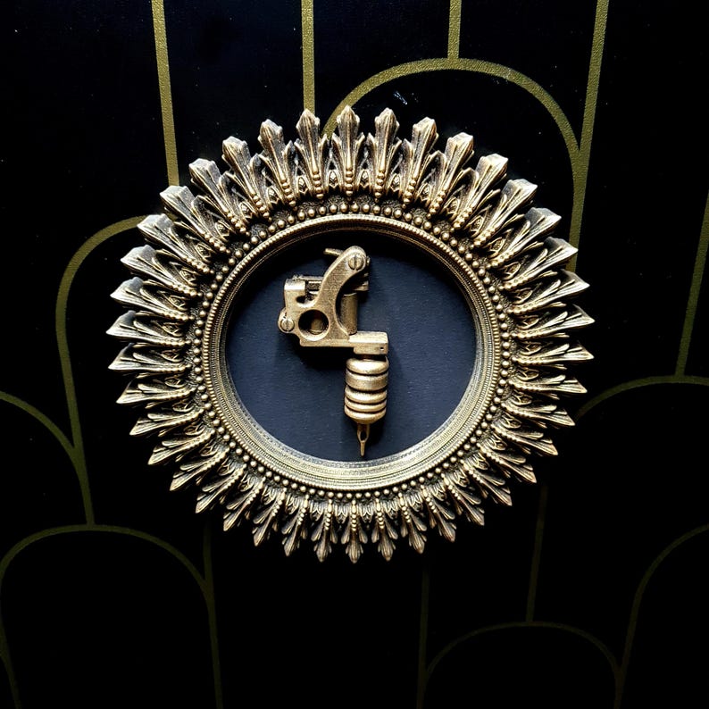 Pode incluir: Uma m&aacute;quina de tatuagem dourada &eacute; exibida em uma moldura circular com um design de raios de sol. A moldura &eacute; feita de metal e tem um fundo preto.