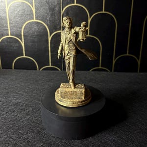 Può includere: Statuetta color bronzo di un uomo in abito che tiene una lanterna. La figura è su una base decorativa sopra una piattaforma circolare nera. Lo sfondo presenta una parete nera con un motivo art déco dorato.
