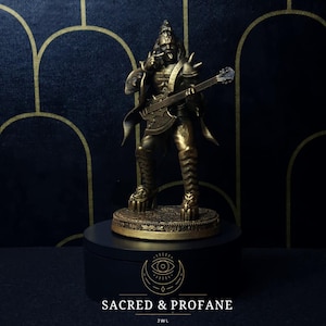 Puede incluir: Una figurita dorada de una persona tocando una guitarra, de pie sobre una base negra con un diseño dorado. La base tiene el texto "SACRED & PROFANE" y "JWL".