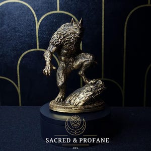 May include: A gold-painted figurine of a werewolf standing on a rock. The figurine is on a black circular base with the text "SACRED & PROFANE" and "JWL" printed on it.
