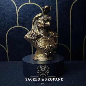 May include: Gold-colored bust sculpture of an Egyptian god with a falcon head, wearing a crown and a necklace. The bust is on a black circular base with a gold logo that says "SACRED & PROFANE" and "JWL".