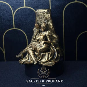 May include: A gold-colored statue of a woman sitting on a throne. The throne is decorated with a Celtic knot design. The woman is wearing a crown and a long robe. The statue is on a black base with the words "Sacred & Profane" and "JWL" written on it.