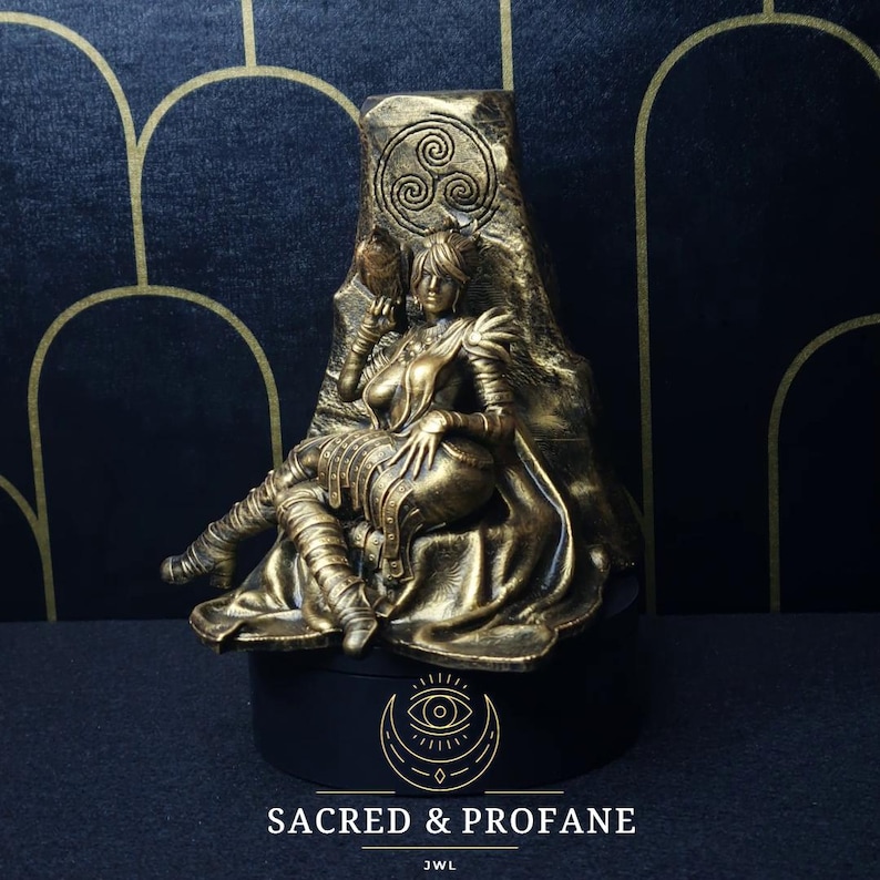 May include: A gold-colored statue of a woman sitting on a throne. The throne is decorated with a Celtic knot design. The woman is wearing a crown and a long robe. The statue is on a black base with the words "Sacred & Profane" and "JWL" written on it.