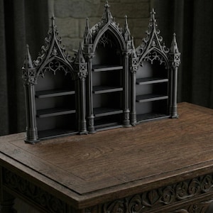 May include: A black, gothic-style bookshelf with three arched sections and multiple shelves. The bookshelf is made of a dark material and sits on a wooden table. The design features intricate architectural details.