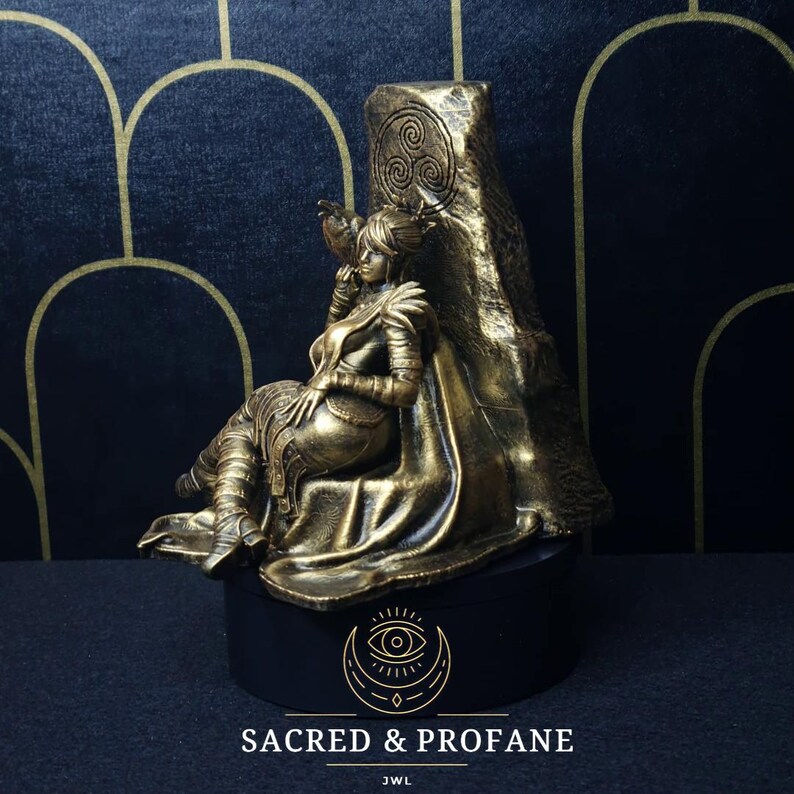 May include: A gold-toned figurine of a woman sitting on a throne. The throne is made of stone and has a Celtic knot design. The woman is wearing a flowing robe and has a crown on her head. The figurine is on a black base with the words "Sacred & Profane" and "JWL" written on it.