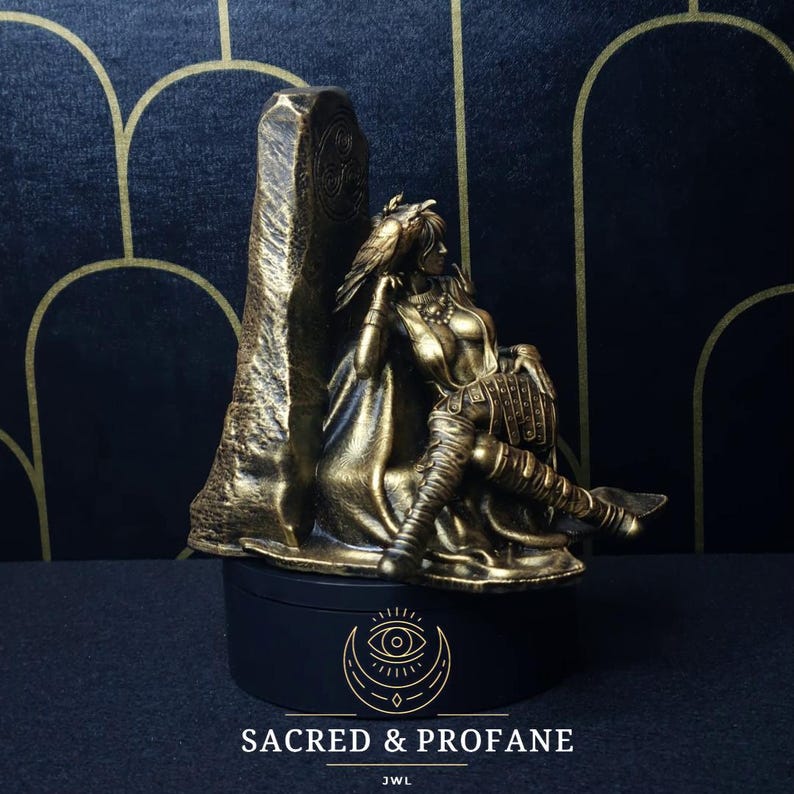 May include: A gold-colored statue of a woman sitting on a throne with a raven on her shoulder. The statue is on a black base with the words "SACRED & PROFANE" and "JWL" written on it.
