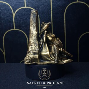 May include: Gold-toned statue of a woman sitting on a throne with a bird on her shoulder. The statue is on a black base with the words "Sacred & Profane" and "JWL" written on it.