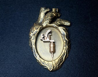 Pode incluir: Um ornamento em forma de coração dourado com um design detalhado. Dentro do coração há uma pequena máquina de tatuagem dourada.