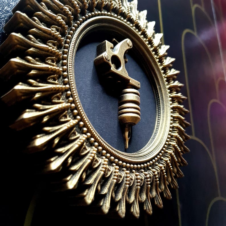 Pode incluir: Uma moldura circular adornada, em tom dourado, com um design detalhado e intrincado. A moldura envolve um fundo preto com uma m&aacute;quina de tatuagem dourada no centro.
