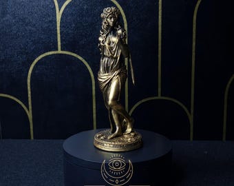 May include: A gold-toned statue of a woman with a floral crown, standing on a black circular base with the text "SACRED & PROFANE" and "JWL".