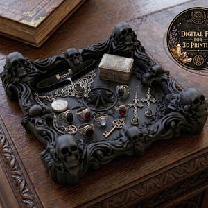 Può includere: Un vassoio portagioie nero in stile gotico con dettagli a teschio, contenente vari anelli, orecchini, una collana e una piccola scatola. Il vassoio è su una superficie di legno. Viene pubblicizzato un file digitale per la stampa 3D.