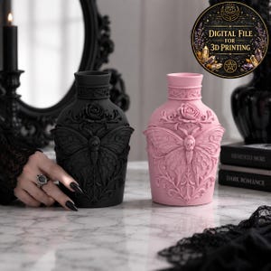 May include: Two decorative vases, one matte black and one pastel pink, each featuring a relief design of a moth with a skull-like thorax, a rose at the neck, and intricate scrollwork. The vases have a cylindrical neck and a rounded body.