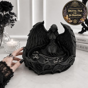 May include: A black circular jewelry tray featuring a central sculpted figure of a winged angel. The tray has ornate, swirling edges and holds two rings and a necklace with a sun-shaped pendant. A circular badge in the corner reads Digital File for 3D Printing.