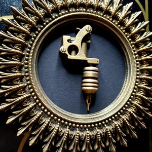 Pode incluir: Uma m&aacute;quina de tatuagem dourada &eacute; exibida em uma moldura circular, ornamentada, de cor dourada. A moldura tem um design de sol com um fundo preto.