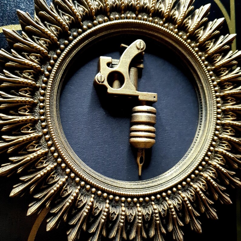 Pode incluir: Uma m&aacute;quina de tatuagem dourada &eacute; exibida em uma moldura circular, ornamentada, de cor dourada. A moldura tem um design de sol com um fundo preto.