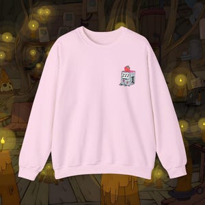 May include: A pale pink sweatshirt featuring a small graphic on the chest. The graphic depicts a cartoon box with the word "ZZZ" and a strawberry. The sweatshirt has a crew neck and long sleeves, suitable for cooler weather.