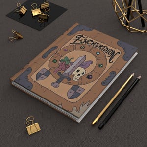 May include: A hardcover journal with the title "Enchiridion" on the cover. The cover features a sword, skull, grapes, and a bird. The journal is brown with a white interior and is surrounded by gold binder clips and pencils.