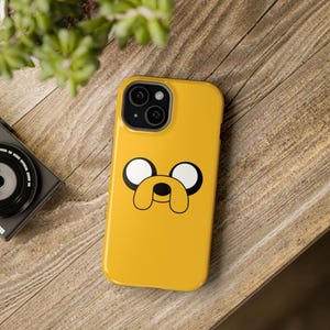 Puede incluir: Funda de teléfono amarilla con un diseño de cara de perro de dibujos animados, con ojos blancos y contornos negros. La funda está colocada sobre una superficie de madera, con una cámara y una planta visibles.