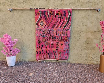 May include: A vibrant pink and multi-colored rug with geometric patterns hangs on a textured wall. The rug features a variety of shapes and colors, including diamonds, triangles, and lines. The rug is suspended from a wooden rod.