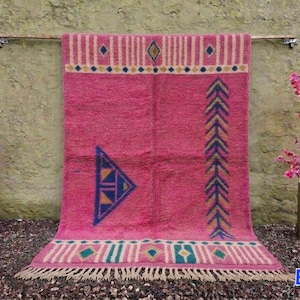 May include: A rectangular rug in a vibrant pink hue featuring a central geometric pattern with a blue triangle and a vertical chevron design. The rug includes horizontal striped borders with small diamond shapes and a fringe along the bottom edge.