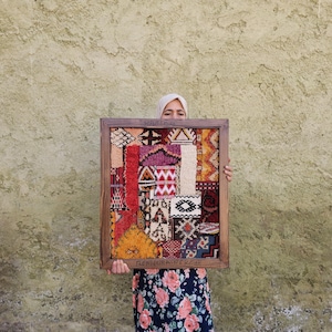 May include: A rectangular wooden frame containing a multi-coloured textile collage with various geometric patterns in shades of red, orange, yellow, black, and white. The bottom edge of the frame features the text "BENTOURA BERBER".