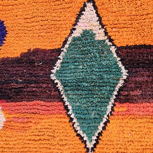 May include: Close-up of a textured rug with a geometric pattern. The rug features a diamond shape in green, outlined in black and white, set against a backdrop of orange, purple, and red hues. The rug's texture is visible.