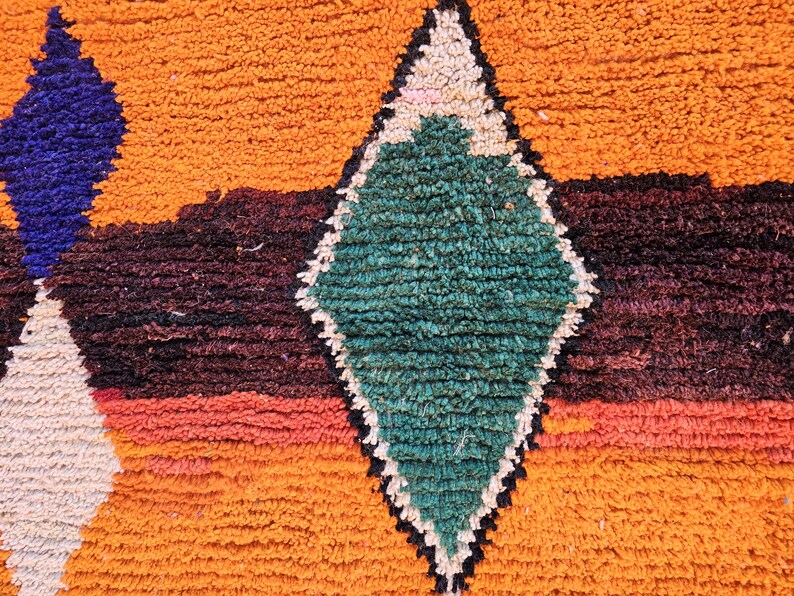 May include: Close-up of a textured rug with a geometric pattern. The rug features a diamond shape in green, outlined in black and white, set against a backdrop of orange, purple, and red hues. The rug's texture is visible.