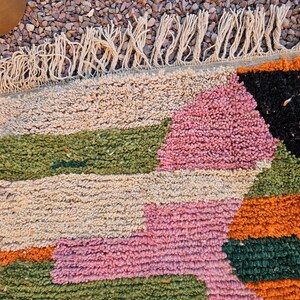 May include: Close-up of a colorful, textured rug with a geometric pattern. The rug features sections of beige, pink, green, orange, and black. The rug has a fringe along one edge, adding a decorative element to the design.