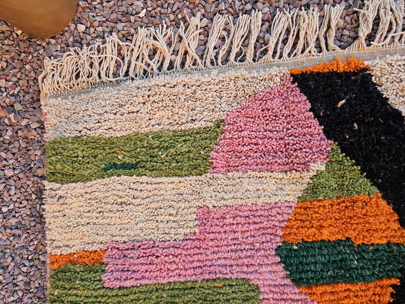 May include: Close-up of a colorful, textured rug with a geometric pattern. The rug features sections of beige, pink, green, orange, and black. The rug has a fringe along one edge, adding a decorative element to the design.