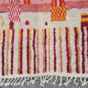Op de afbeelding: Een close-up van een kleurrijk, getextureerd tapijt met een cr&egrave;mekleurige basis. Het tapijt heeft verticale strepen in tinten rood, roze en geel, met geometrische patronen in het bovenste gedeelte. Het tapijt heeft een gefranjerde rand.