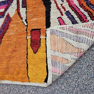 May include: A colorful, textured rug with a geometric design. The rug features shades of orange, yellow, red, pink, purple, and white. The rug is folded over, revealing the underside with a striped pattern.