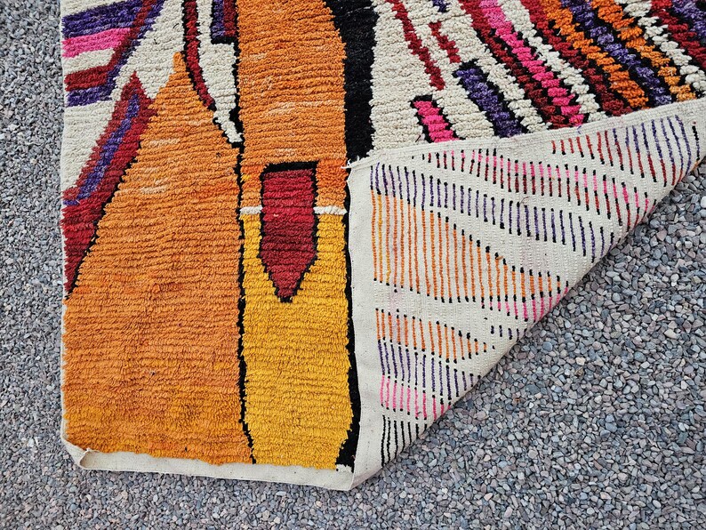 May include: A colorful, textured rug with a geometric design. The rug features shades of orange, yellow, red, pink, purple, and white. The rug is folded over, revealing the underside with a striped pattern.