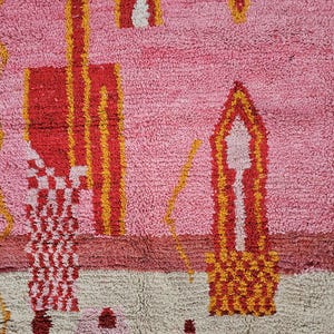 Op de afbeelding: Een close-up van een roze, rood en geel patroonkleed. Het kleed heeft geometrische vormen en ontwerpen in tinten roze, rood, geel en cr&egrave;me. Het kleed heeft een getextureerde, geweven uitstraling.