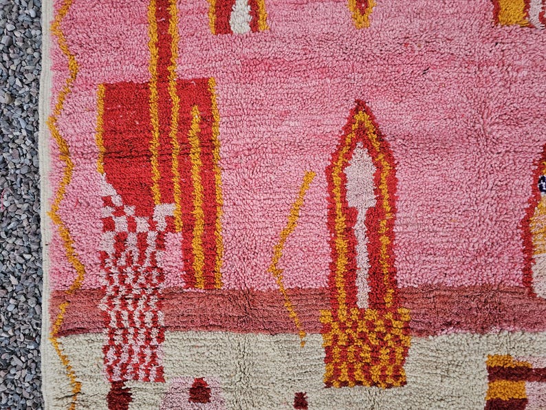 Op de afbeelding: Een close-up van een roze, rood en geel patroonkleed. Het kleed heeft geometrische vormen en ontwerpen in tinten roze, rood, geel en cr&egrave;me. Het kleed heeft een getextureerde, geweven uitstraling.