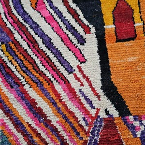 May include: Close-up of a colorful, textured rug with a geometric design. The rug features diagonal stripes in shades of pink, purple, orange, and red against a white background. A section of the rug has a black and orange pattern.