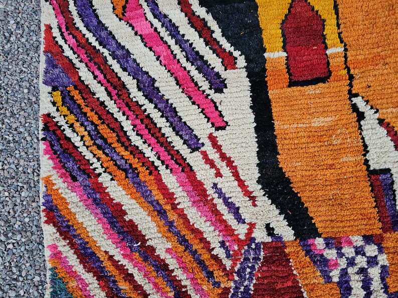May include: Close-up of a colorful, textured rug with a geometric design. The rug features diagonal stripes in shades of pink, purple, orange, and red against a white background. A section of the rug has a black and orange pattern.